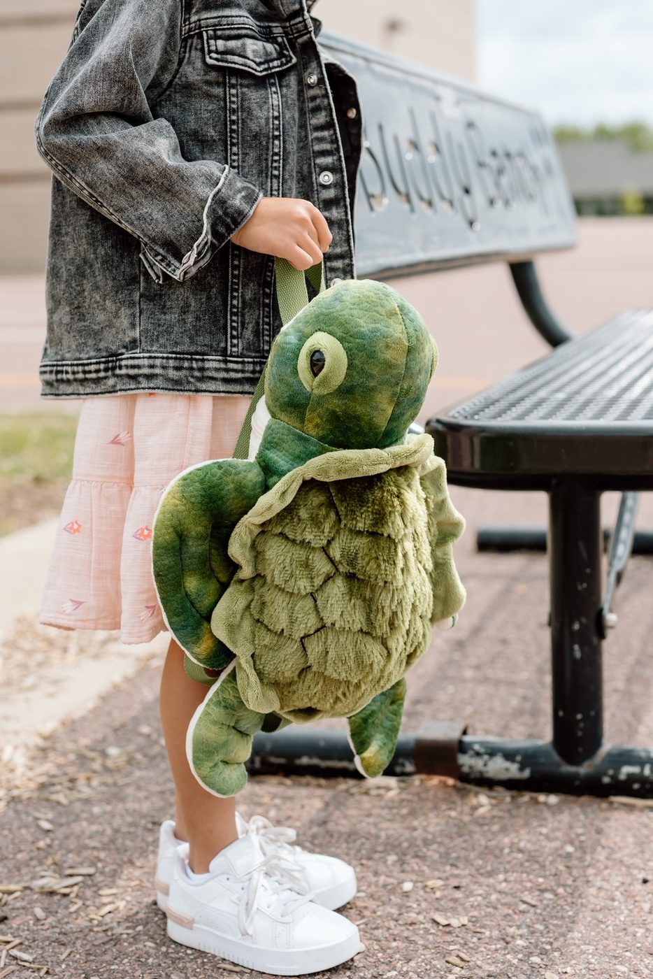 A child outdoors wearing a green turtle plush backpack